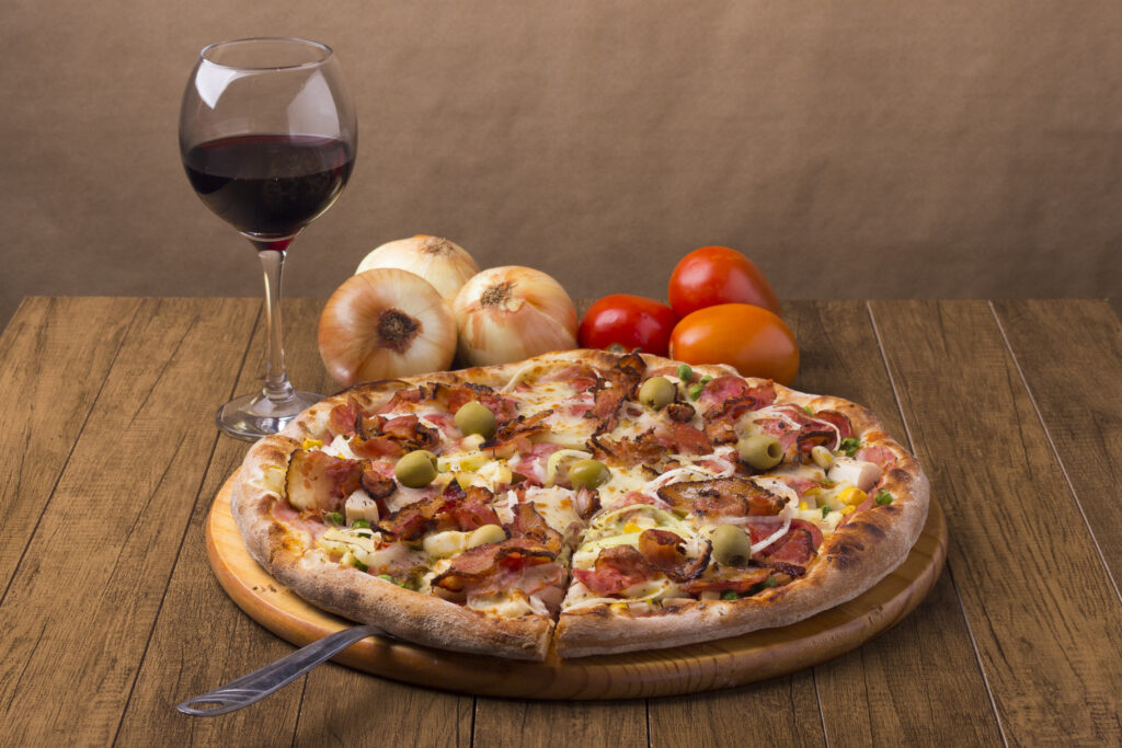 Tasty Portuguese Pizza made with ham, pea egg, heart of palm, pepperoni, onion and mozzarella and bacon. Glass of Red Wine. Slice of pizza ready to serve. Gastronomy photo of pizzas.